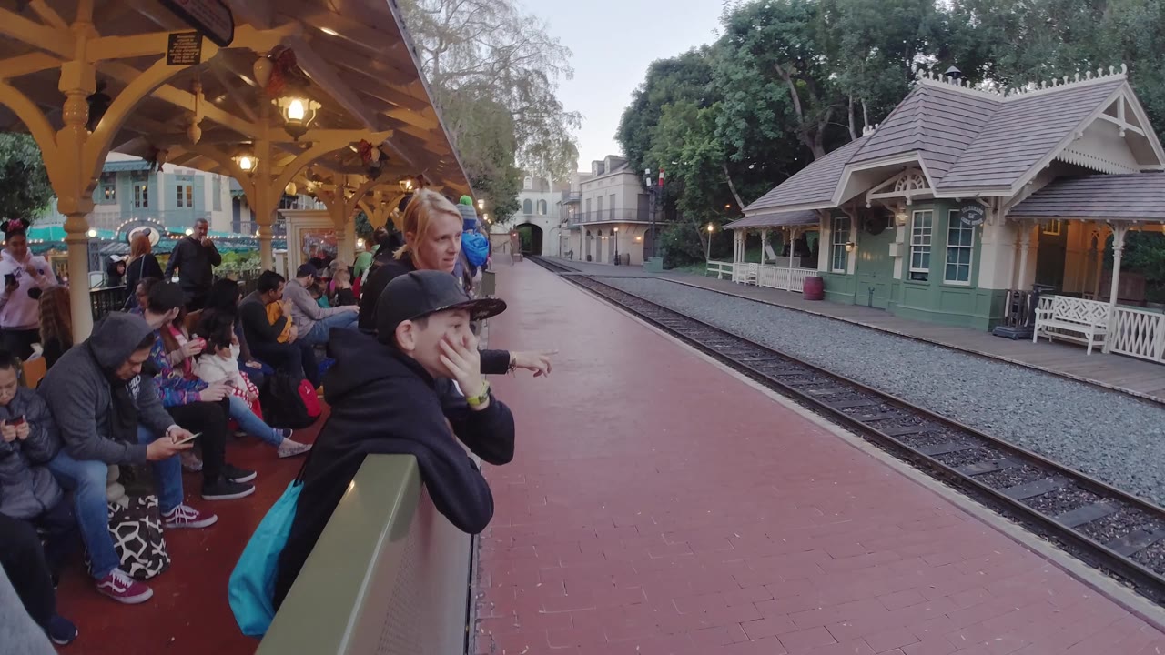 At Disneyland, California, a boy in a dark cap leans in the foreground as other guests wait under the station roof beside the empty rails.