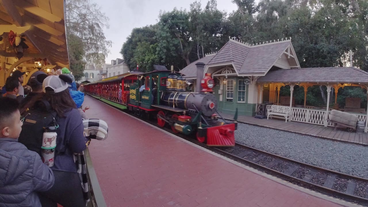 At Disneyland California, a colorful amusement park train approaches the platform while adults and children wait under a covered area beside the track.