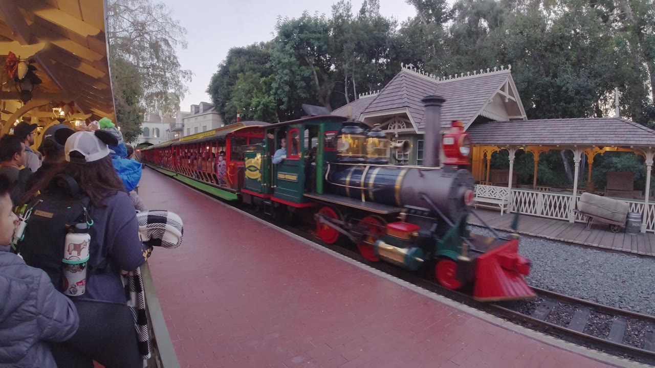 At Disneyland California, the engine passes close to the platform camera, with red wheels, a dark boiler, green trim, and nearby guests partly framing the left side.