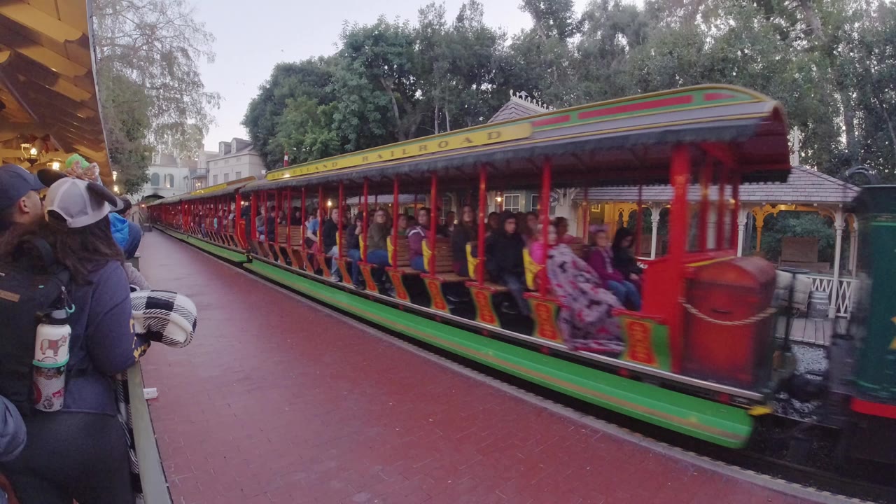A red, green, and yellow Disneyland California train passes beside a red walkway, with riders in the open cars and bystanders partly visible at the left edge.
