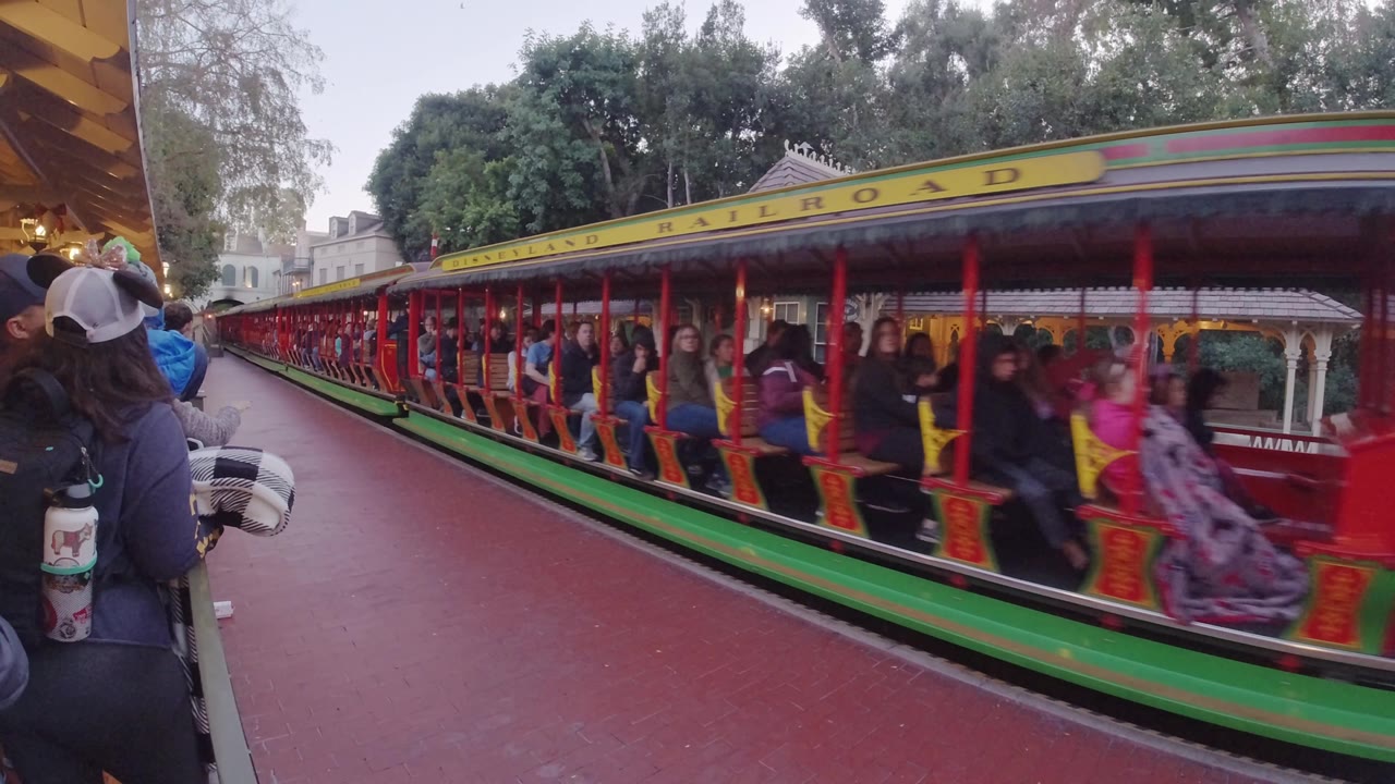 The train advances through the frame with more riders visible under the yellow Disneyland Railroad roof sign, while the red platform and trees remain in the background.