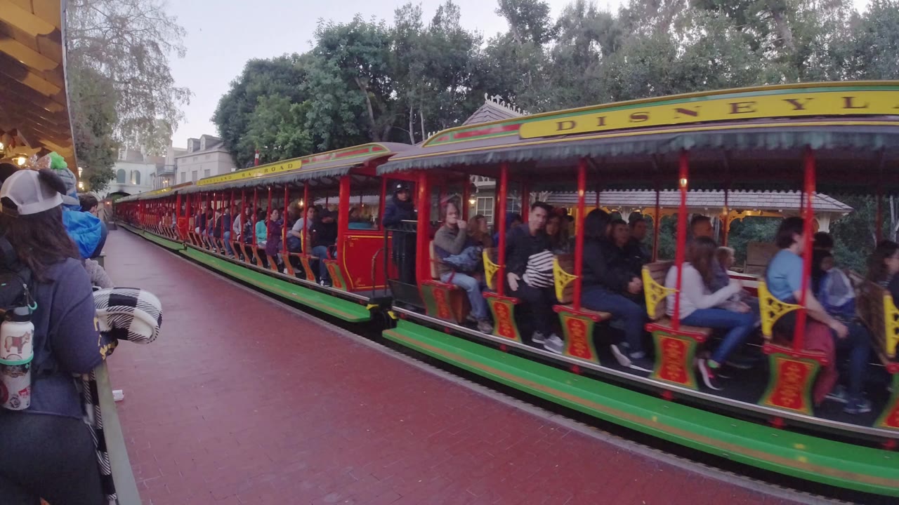 A red Disneyland Railroad train with seated passengers passes close to the platform at Disneyland California, with trees and park buildings behind it.