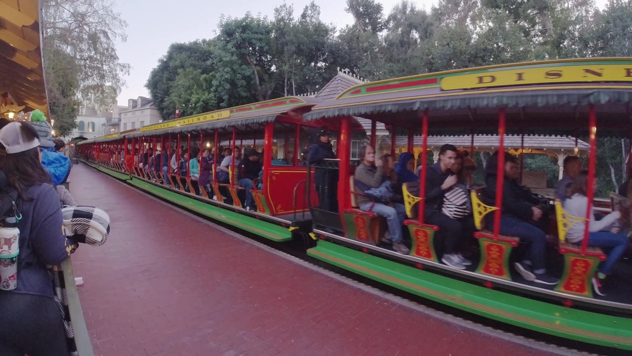 A later moment shows the red and green Disneyland California train shifted along the platform, with passengers in the foreground cars and the yellow roof sign partly cropped.