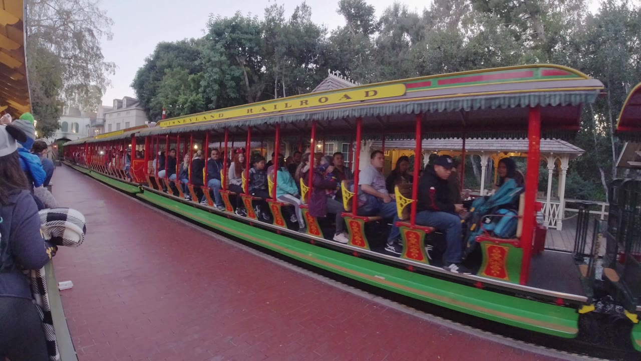 A green, red, and yellow Disneyland Railroad train sits close to the camera in Disneyland California, with passengers visible in open cars and red pavement filling the foreground.