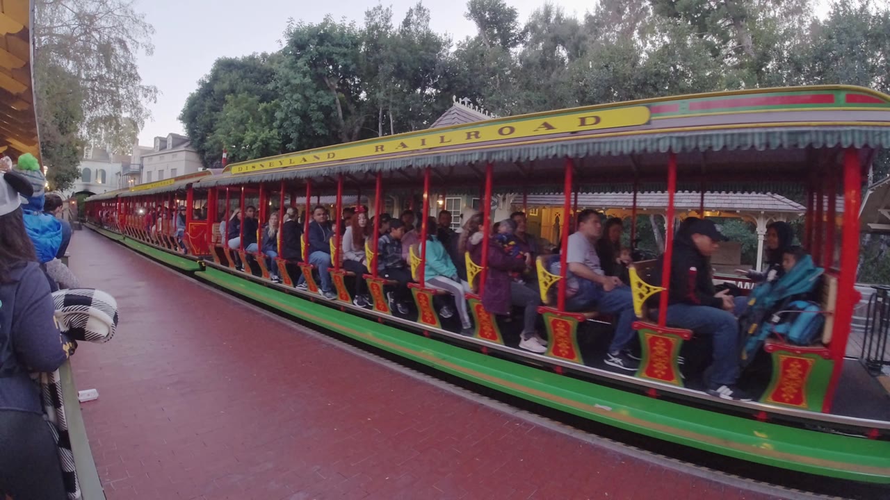 A closer section of the Disneyland Railroad train is visible in Disneyland California, with riders seated under the yellow roof sign and the green lower carriage running across the frame.