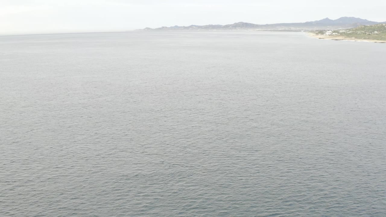 Aerial view in Baja California Sur, Mexico, showing open rippled water, a thin far coast, muted mountains, pale daylight, and no visible people.