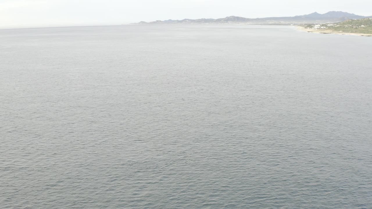 High aerial frame from Baja California Sur, Mexico, with broad gray water in the foreground, distant sandy shoreline, low hills, and no visible people.