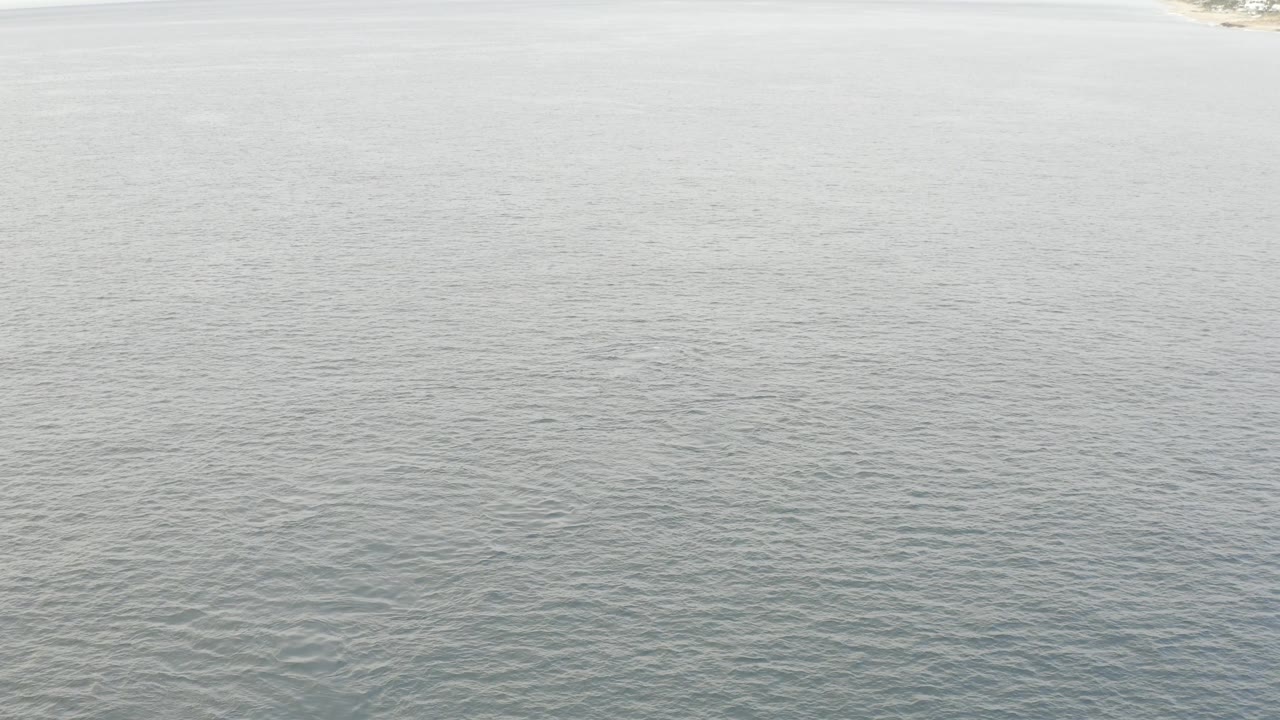 Wide drone-style view across calm grey lake water in Baja California Sur, with a brighter shoreline edge near the top of frame and no visible people.