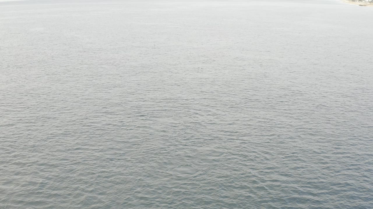 Aerial frame over Baja California Sur water showing rippled blue green ocean, a narrow pale sand edge at the top, and no visible people.