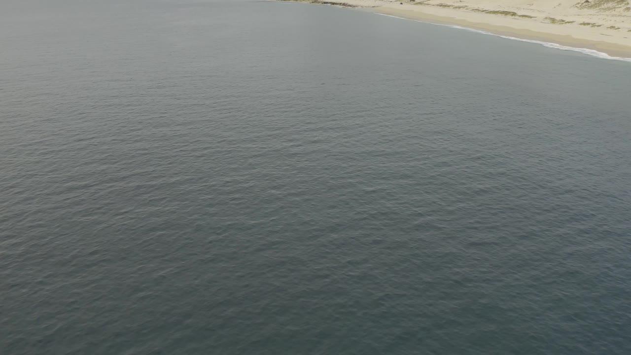 Aerial view across calm blue-green water toward a curving Baja California Sur beach at the top right, with no visible people.