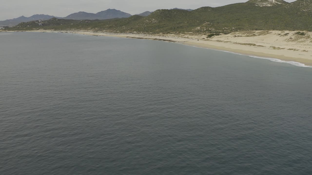 Wide aerial view of Baja California Sur coastline with ocean in front, sandy beach and dunes in the middle, scrub hills and distant mountains behind, and no visible people.