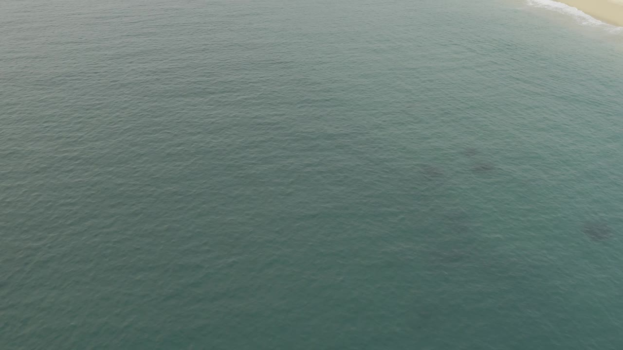 Wide aerial view of calm green-blue ocean off Baja California Sur, with a thin sandy coastline at the upper right and no visible people.