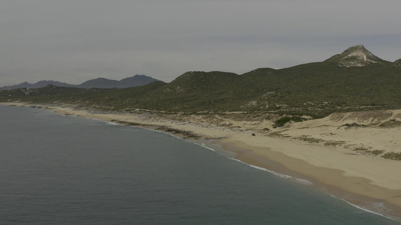 Aerial view over Baja California Sur coastline with blue-green water in the foreground, a pale sand beach curving below green hills, and no visible people.