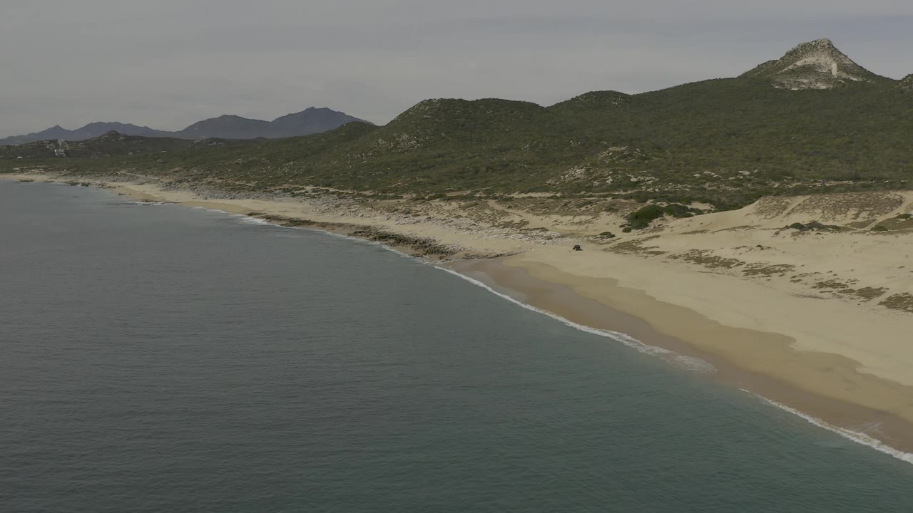 Wide drone-style view of Baja California Sur coastline, showing a broader sweep of water, beach, dunes, green hills, and no visible people.