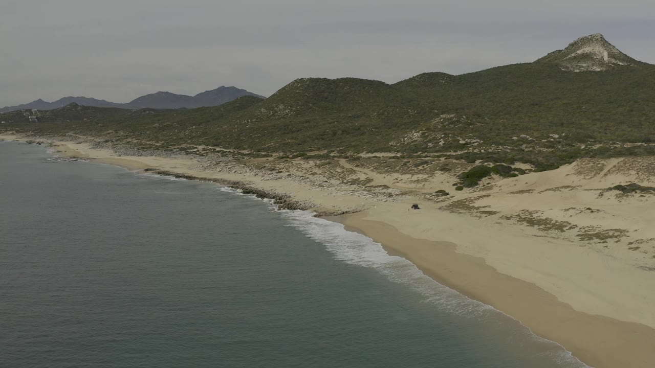 Wide aerial frame of Baja California Sur beach with small white surf lines, dune scrub across the midground, a prominent mountain peak near the upper right, and no visible people.