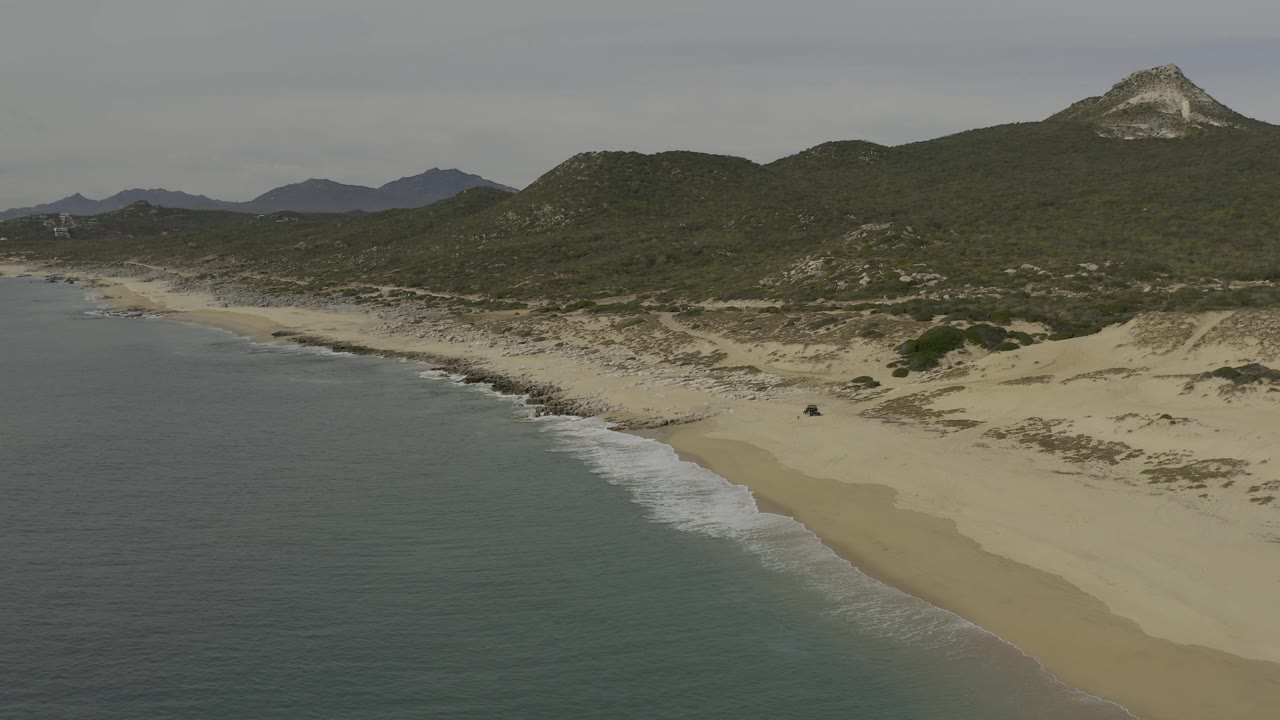 Oblique aerial view along the Baja California Sur shoreline, with muted water on the left, a broad empty beach on the right, green hills and a pale rocky summit in the background, and no visible people.