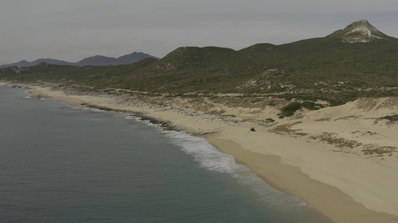 Aerial view over Baja California Sur coast as a low wave line reaches the sand, with scrub-covered dunes leading to layered hills and no visible people in the frame.