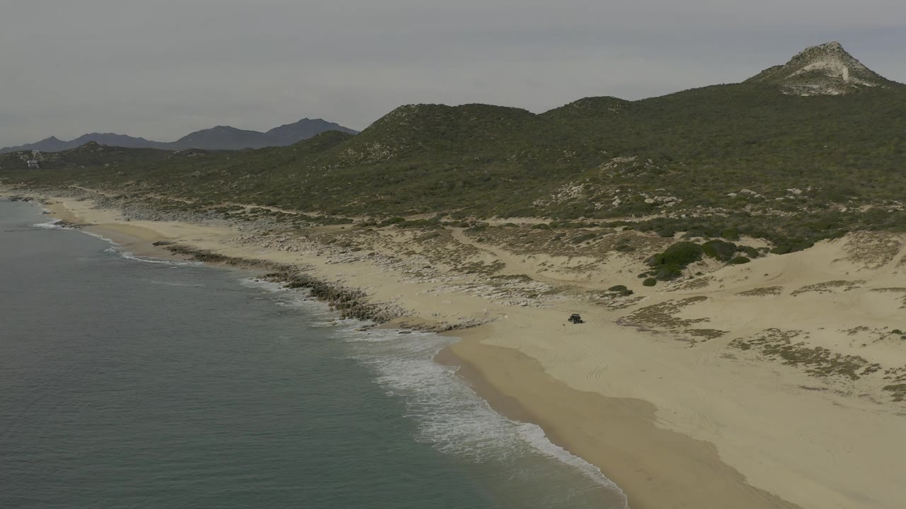 Aerial frame at 0:01 shows the Baja California Sur shoreline curving below the drone, with surf on the left, sand flats in front, dark vegetation on the hills, and no visible people.