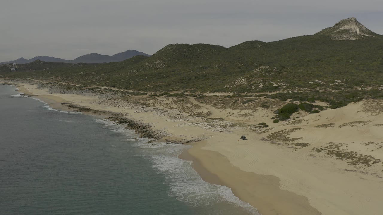 At 0:02 the aerial view keeps the ocean on the left while a broad sandy beach and dune field fill the lower frame, with Baja California Sur hills and mountains beyond and no visible people.