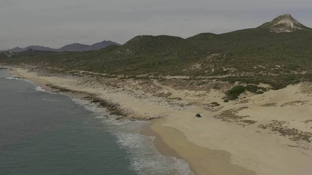 Frame 0:03 shows the Baja California Sur coast from above, with foamy waves along the left edge, rocky sand and dunes below, dark green hillside vegetation, distant peaks, and no visible people.