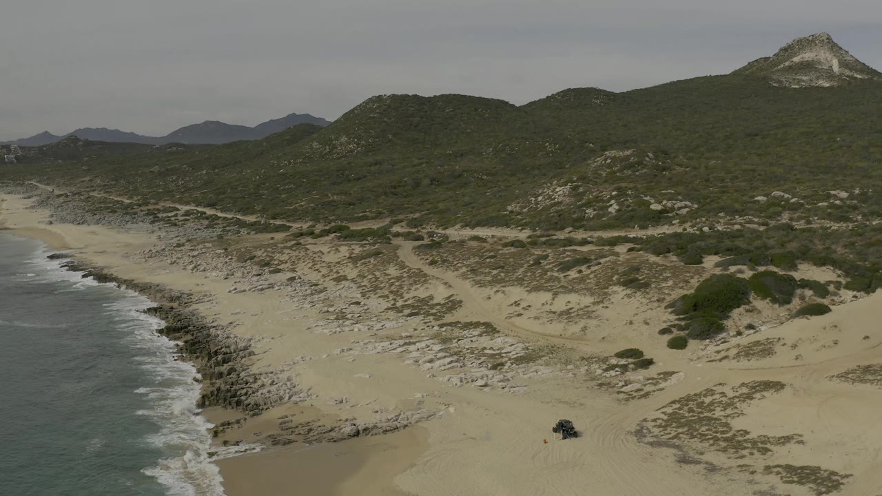 Aerial frame over Baja California Sur coastline with gray-green surf at left, pale sand and dunes across the center, green hills in back, a dark vehicle on the beach, and no visible people.