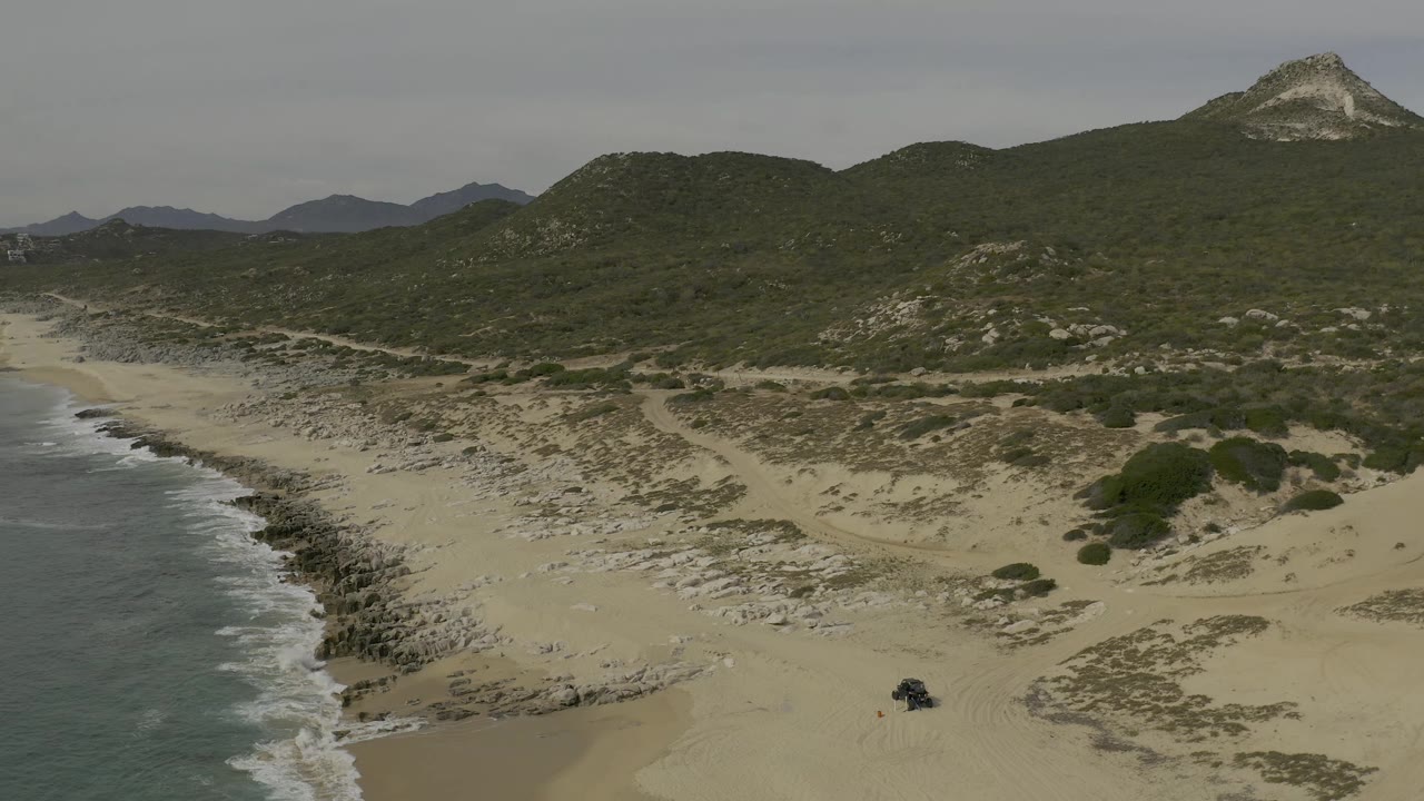 Second aerial frame over Baja California Sur with the surf band shifted slightly along the left edge, open sand and dune tracks visible below, hills behind, a dark vehicle on the lower beach, and no visible people.