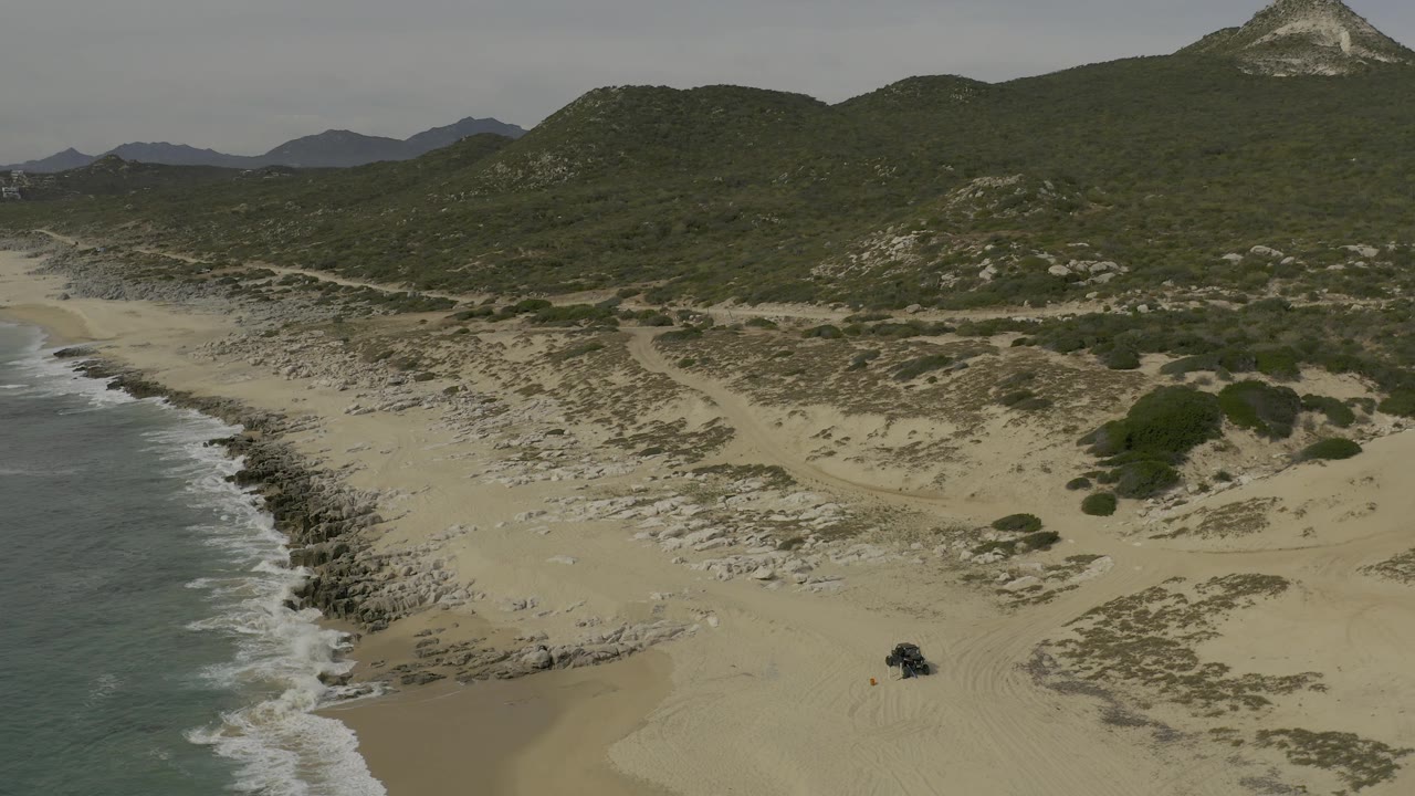 Aerial view in Baja California Sur showing more of the curved beach and rocky waterline at left, pale dune fields at center, shrub patches at right, distant hills, a dark vehicle, and no visible people.