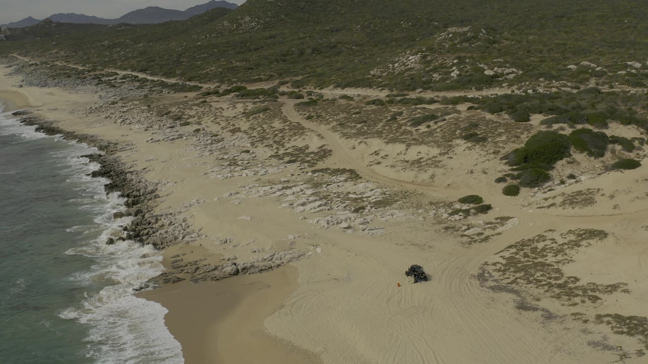 Drone-height view over Baja California Sur beach with foamy waves along the left margin, rock-streaked sand and scrub through the middle, hills across the top, a dark vehicle near the lower right, and no visible people.