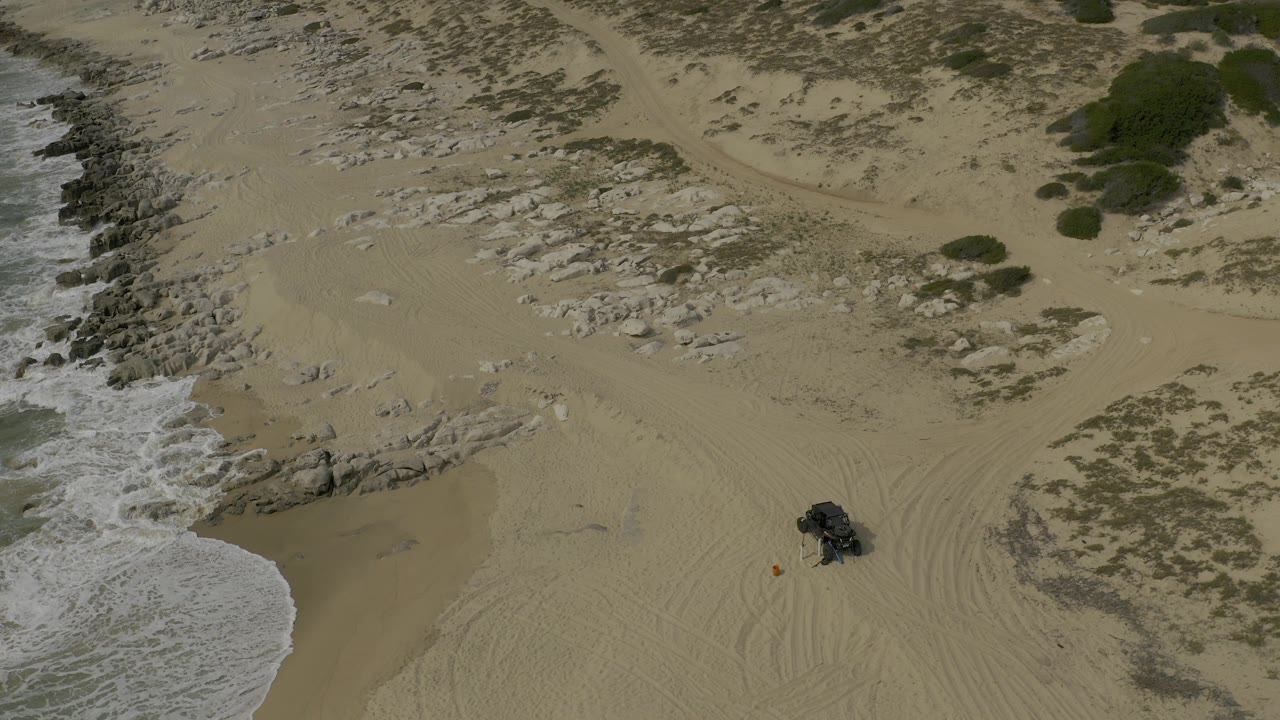 Overhead view of a Baja California Sur beach with white surf at left, pale sand, rock shelves, tire tracks, a dark off-road vehicle near the lower center, and no visible people.