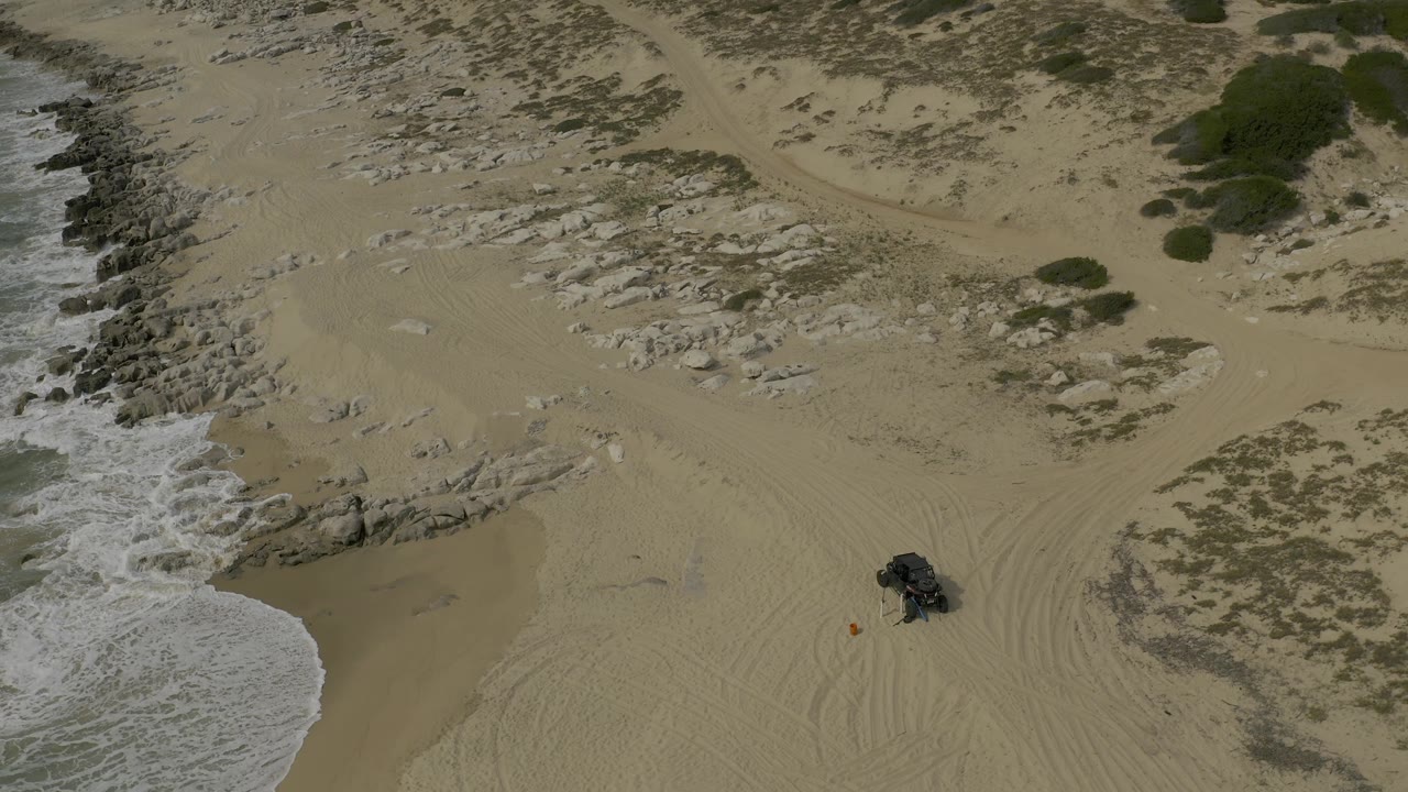 Overhead coastline with waves, rock ledges, sand tracks, and a dark vehicle.