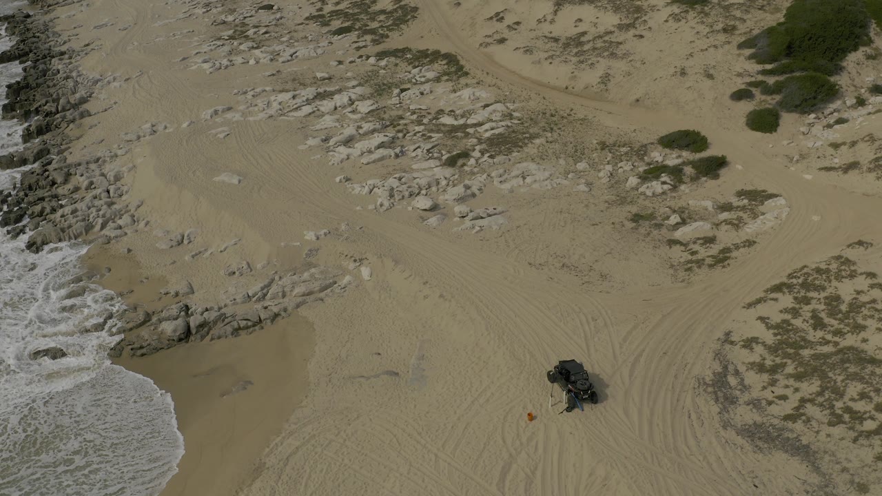 High aerial view in Baja California Sur with the surf line pressing into the left side, pale rock formations across the upper beach, multiple tire marks, a dark off-road vehicle to the lower right, and no visible people.