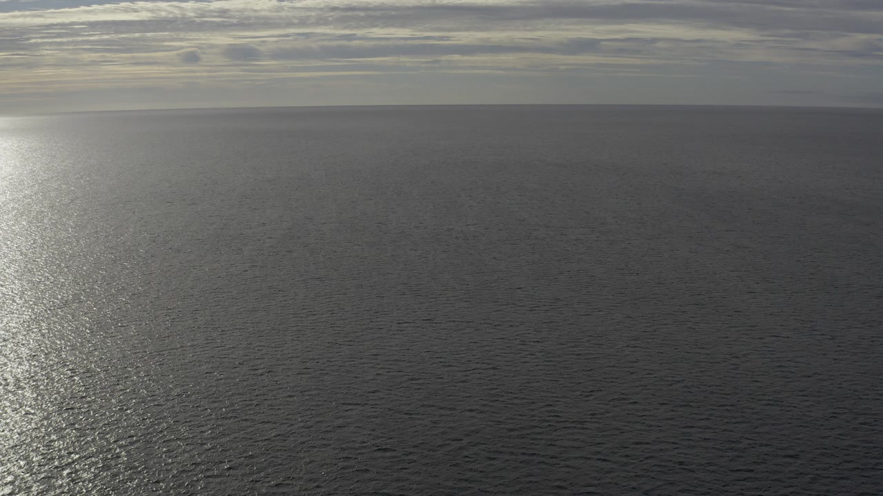 Wide aerial view over Baja California Sur ocean water with a low horizon, layered cloud, bright sun reflection at left, and no visible people.