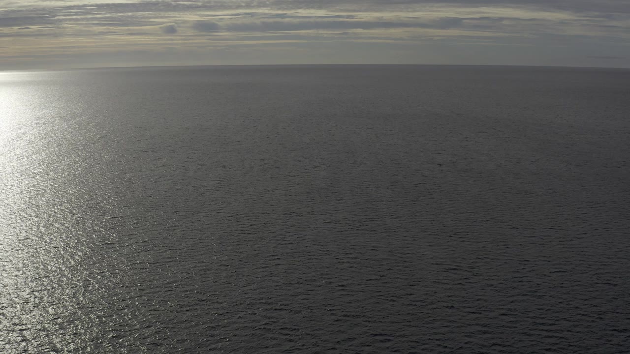 Aerial view over Baja California Sur ocean water at sunset, with a bright glare path near the left side, layered clouds on the horizon, and no visible people.