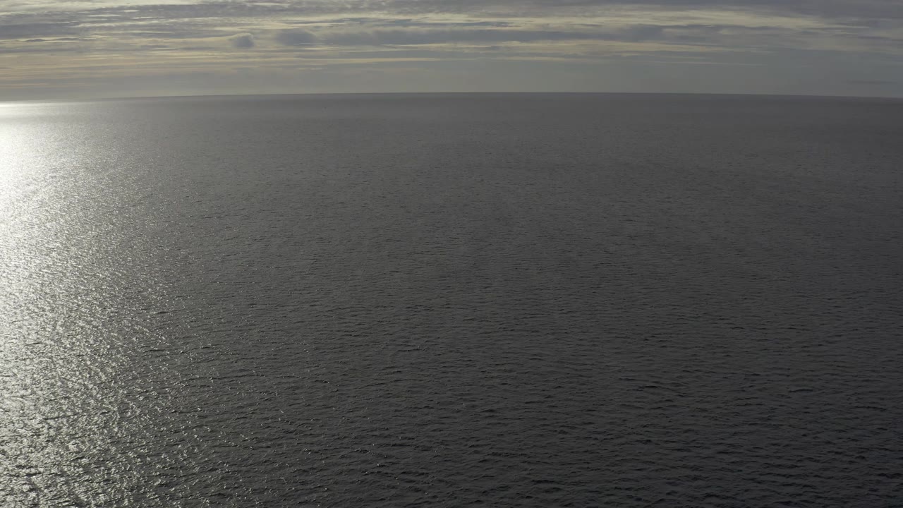 Aerial Baja California Sur ocean frame with the sun glare closer to center-left, dark rippled water below, cloud bands above, and no visible people.