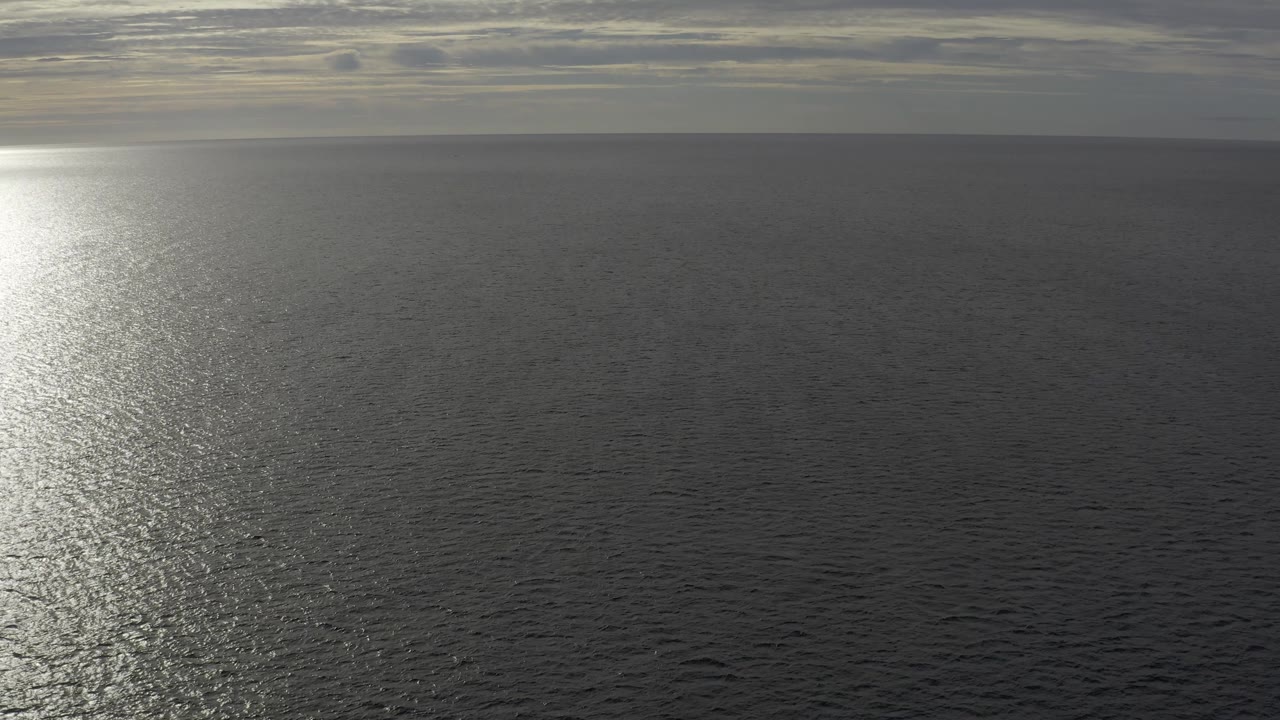 Aerial Baja California Sur seascape with the bright reflection shifted to the far left edge, broad dark water in the foreground, cloud layers at the horizon, and no visible people.