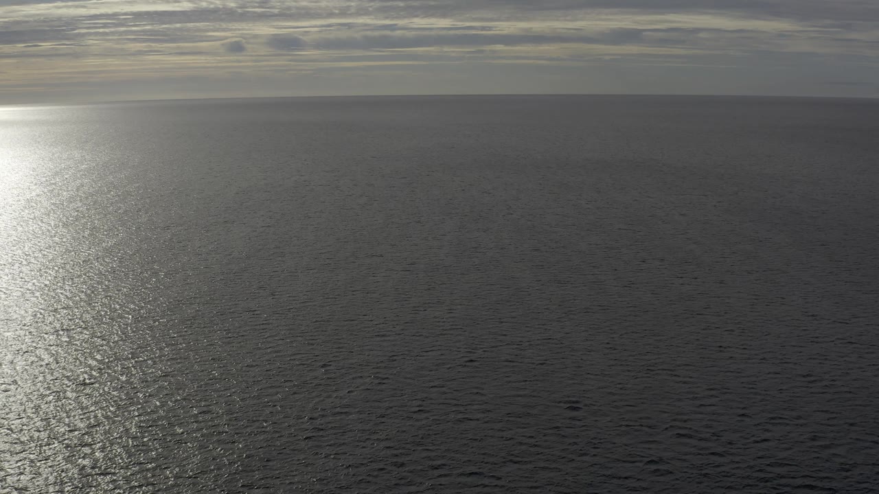Wide ocean horizon in Baja California Sur with the bright reflection pushed toward the left edge, flat gray cloud layers above, textured water below, and no visible people.