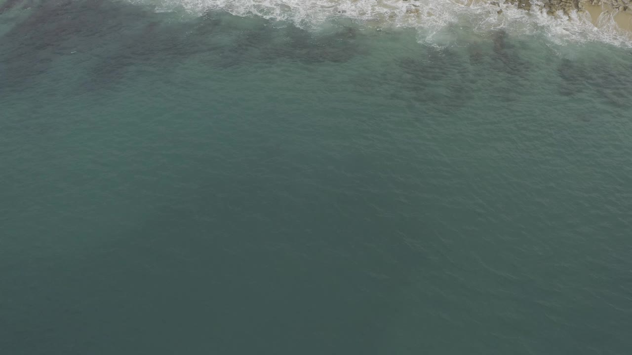 Overhead view of Baja California Sur water with white surf touching the sandy shore at the top edge and no visible people.