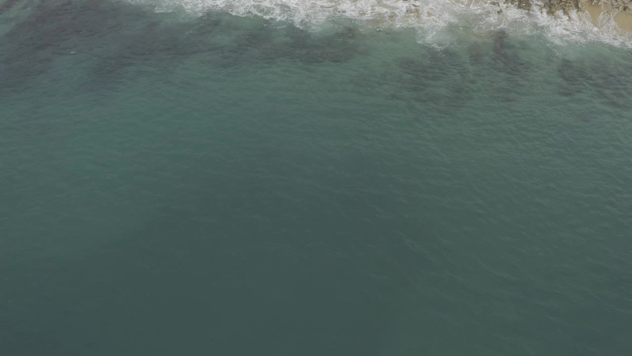 Aerial frame over Baja California Sur coastline, with blue-green water dominating the view and a narrow white wave band along the top, no visible people.