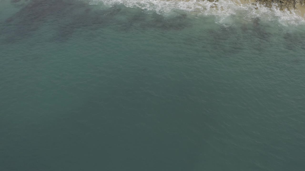 Top-down Baja California Sur shoreline view where foamy surf spreads unevenly across the top edge and textured ocean fills the frame, no visible people.