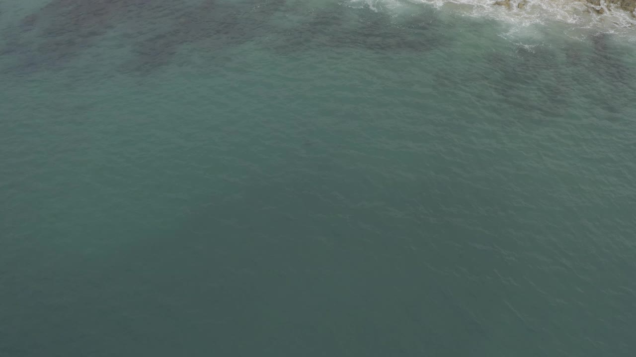 Aerial view in Baja California Sur, Mexico showing rippled green-blue sea and pale surf touching the narrow coast at the top of frame, no visible people.