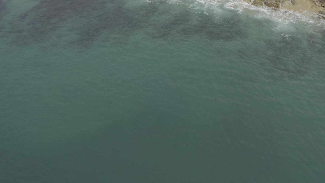 High aerial frame over Baja California Sur, Mexico where open green-blue sea dominates and small broken waves mark the shoreline near the top, no visible people.