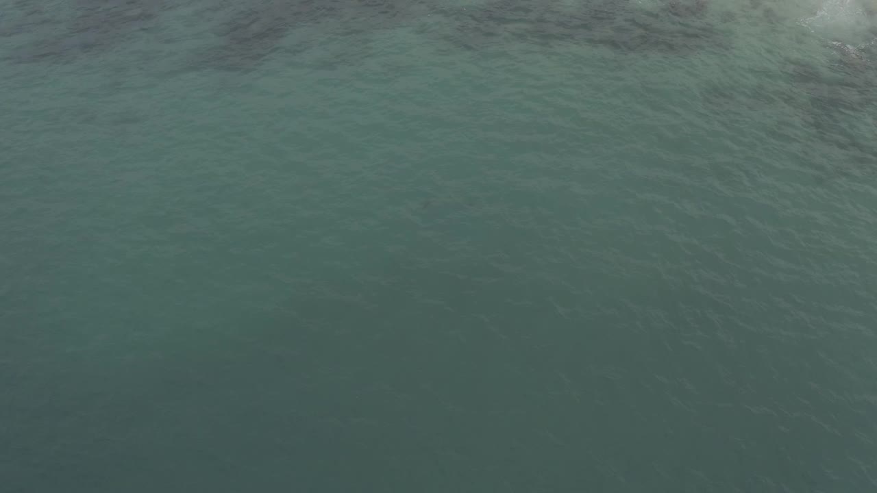 Aerial ocean view in Baja California Sur, Mexico, with darker green water below and pale wave foam entering along the upper edge, with no visible people.