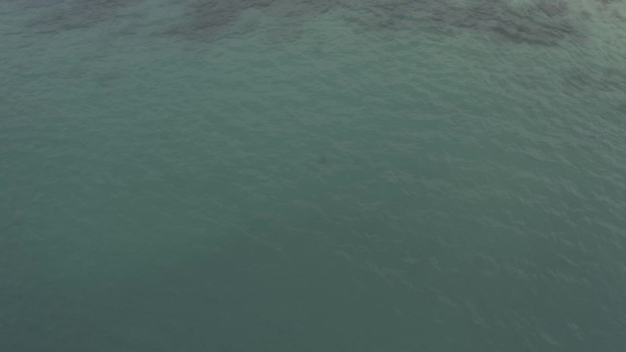 Aerial view above the Baja California Sur sea surface, where pale foam and darker wave shadows run across the upper frame above textured green water, with no visible people.