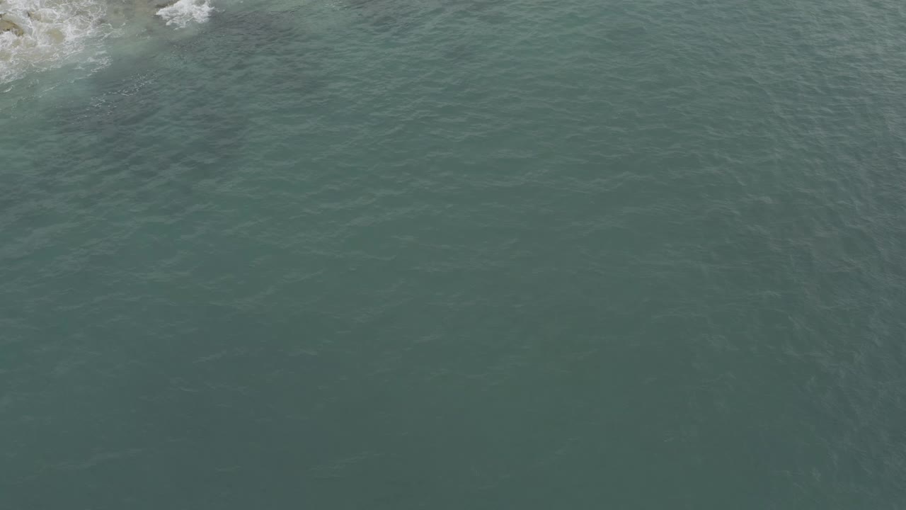 Aerial frame over Baja California Sur water with most of the view occupied by textured green-blue sea and a small foamy wave line touching rocks near the top, no visible people.