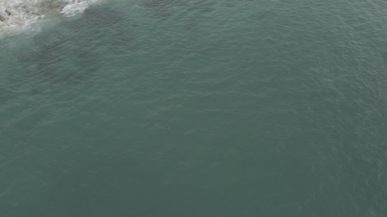 Aerial view in Baja California Sur showing green coastal water, faint current lines, and a small patch of white surf against grey rocks at the upper edge, no visible people.