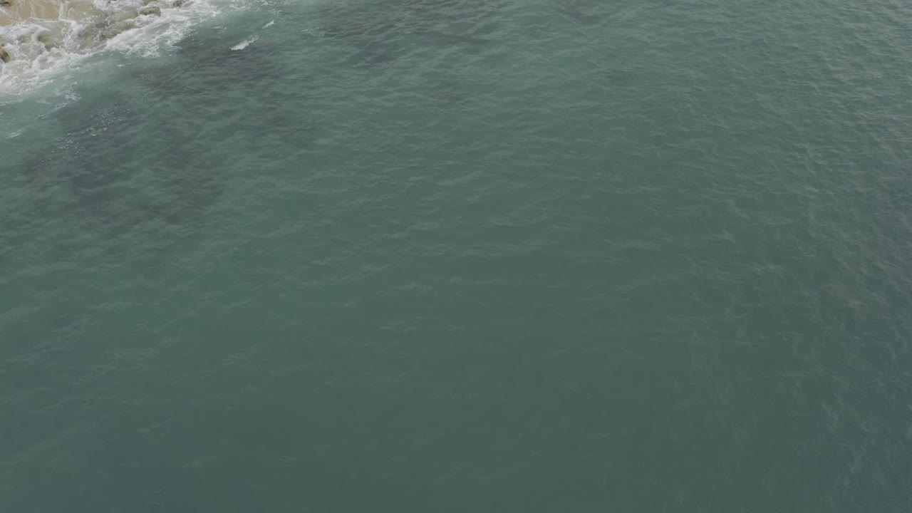 Aerial frame from Baja California Sur with a rocky corner entering from the upper left, small white waves curling around it, and muted green water across the rest of the image, no visible people.