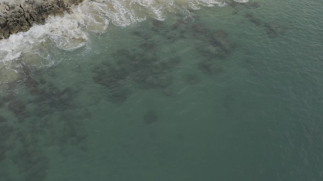 Overhead Baja California Sur coastline frame with white foam curling around gray rocks, muted green shallows, and darker underwater patches, with no visible people.