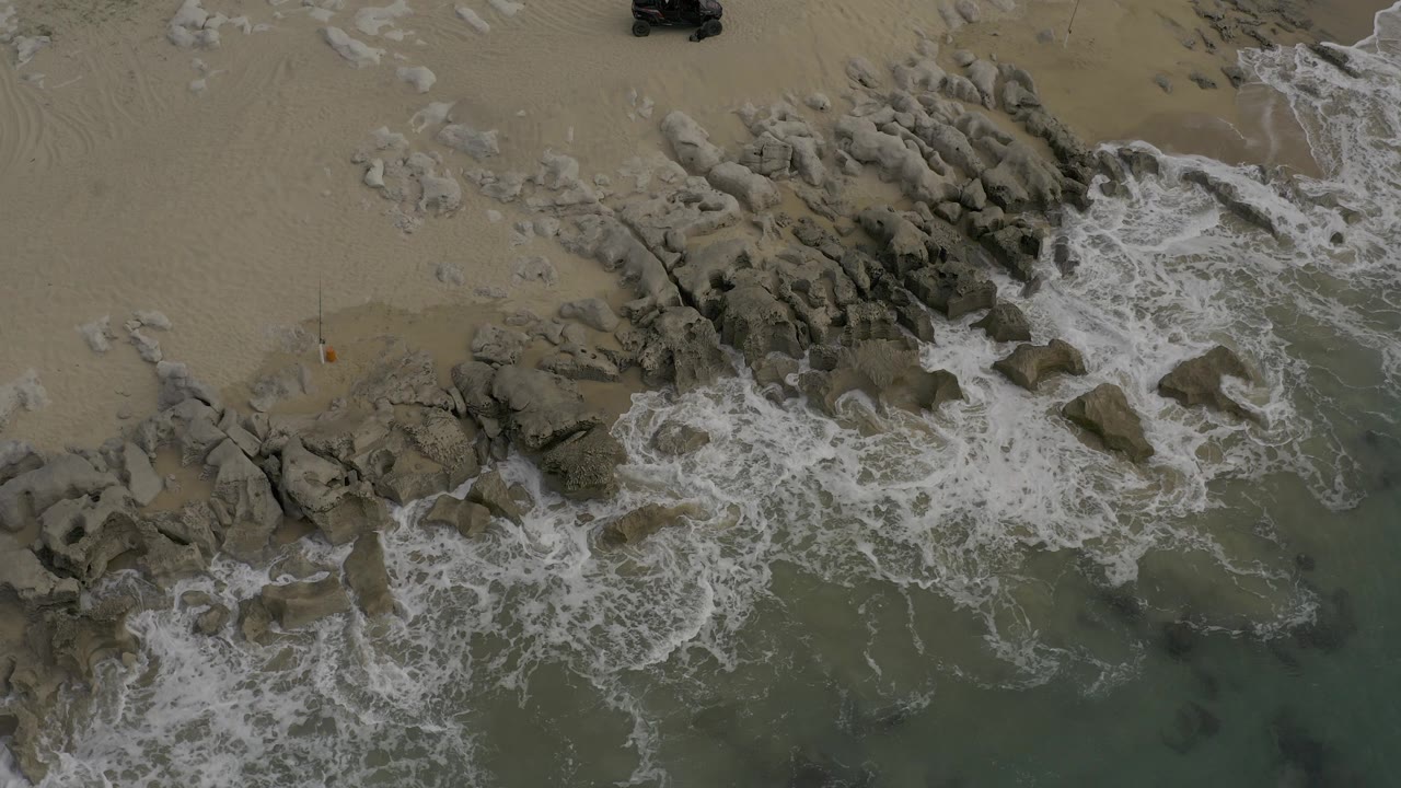 Aerial view of white surf breaking around rocks beside sand.