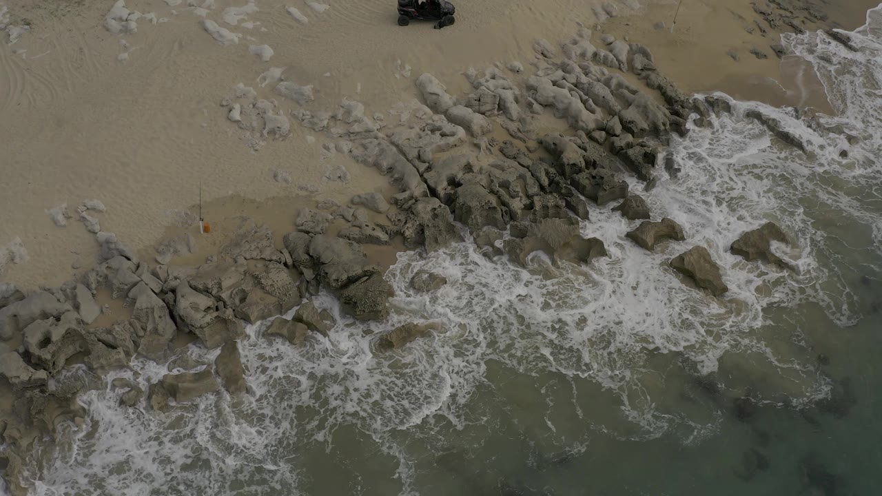 Overhead frame from Baja California Sur, Mexico, showing foam curling around a central rock cluster with open sand above and no visible people.