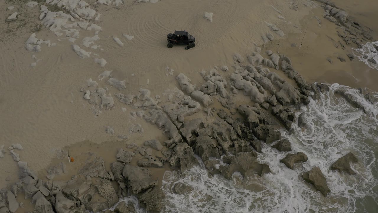 Aerial frame in Baja California Sur, Mexico, with surf hitting rocks at the lower right, broad sand above, a small dark off-road vehicle near the top edge, and no visible people.